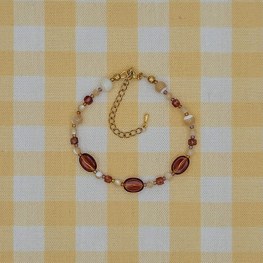 A gold-tone beaded bracelet with oval reddish-brown beads and small pearl-like accents, laid flat on a yellow and white gingham checkered fabric background.