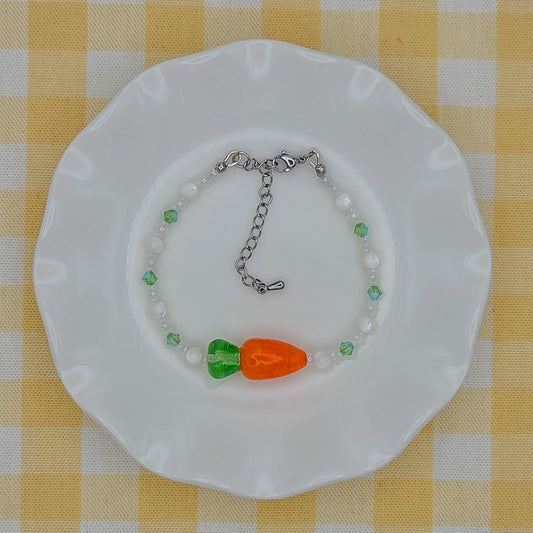A beaded bracelet with white pearls, green beads, and a carrot charm, displayed on a white scalloped plate over a yellow and white checkered tablecloth.