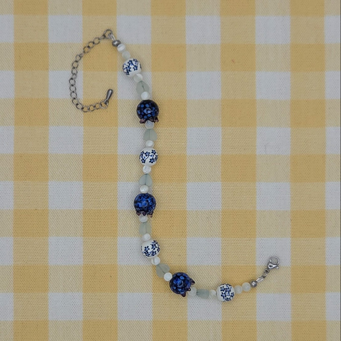 A beaded necklace with blue and white patterned beads and silver-toned chain links, laid flat on a yellow and white checkered fabric background.