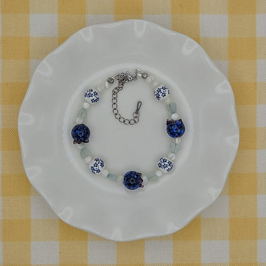 A beaded bracelet with blue and white floral beads and silver-toned chain clasp, displayed on a white scalloped plate over a yellow and white checkered tablecloth.