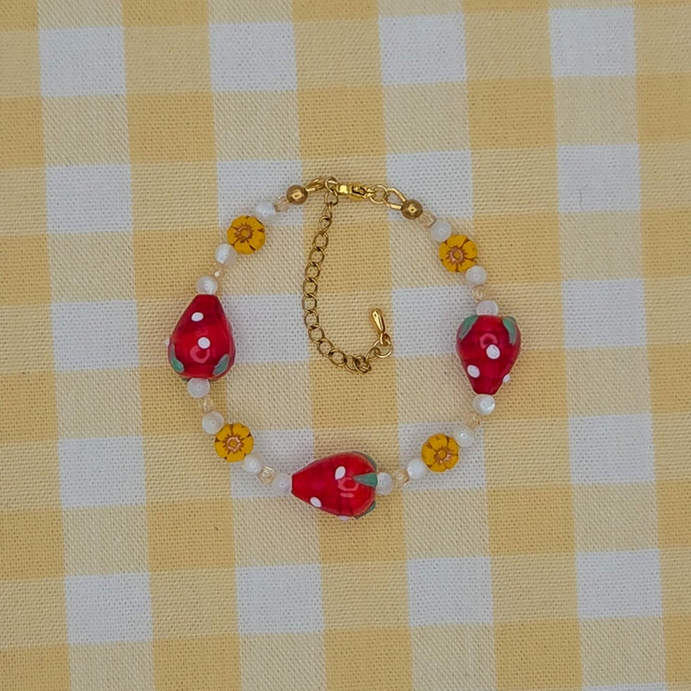 A handmade beaded bracelet featuring red strawberry-shaped beads, white pearl-like beads, and small yellow flower beads on a gold chain, displayed on a yellow and white checkered fabric background.