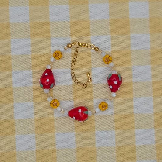A handmade beaded bracelet featuring red strawberry-shaped beads, white pearl-like beads, and small yellow flower beads on a gold chain, displayed on a yellow and white checkered fabric background.