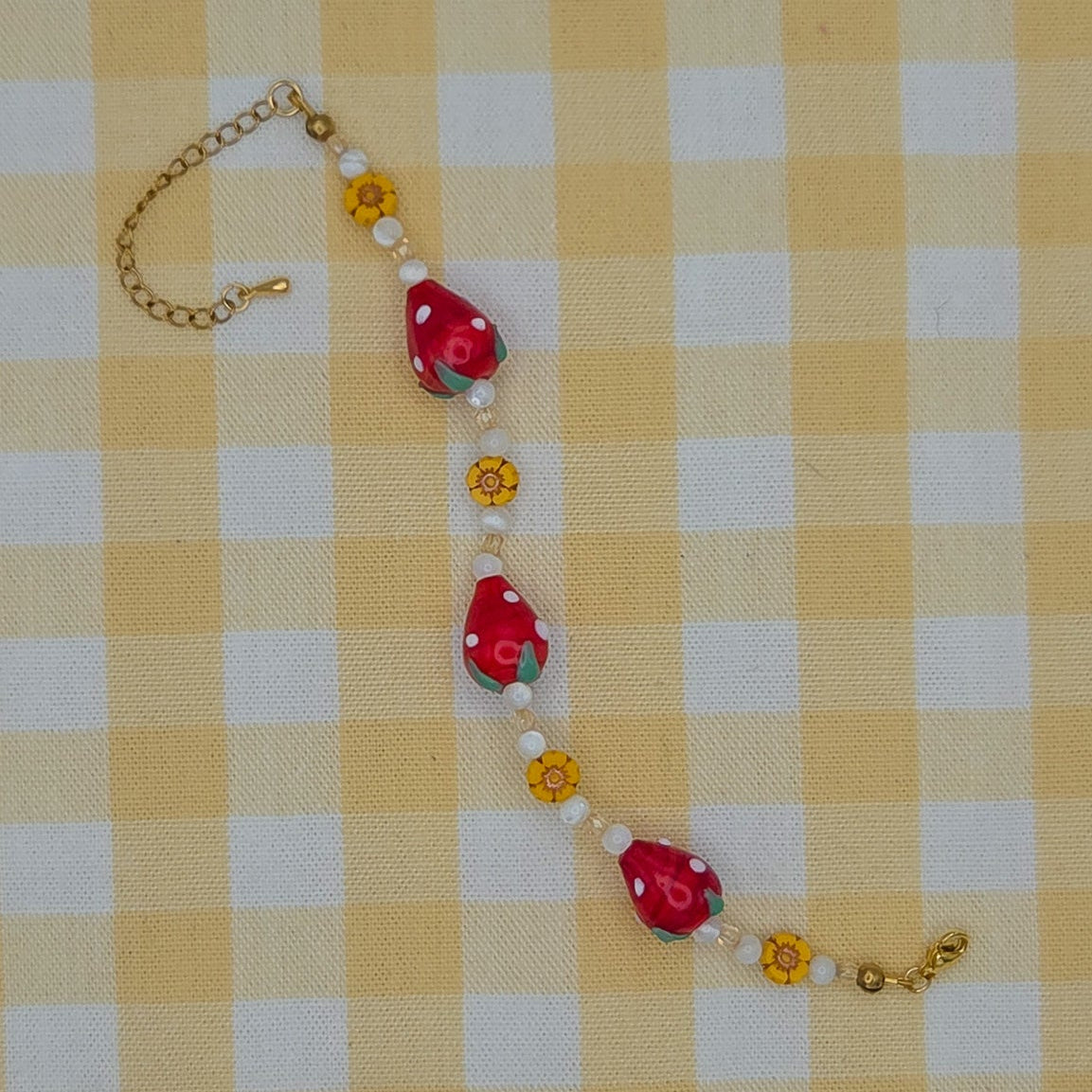 A handmade beaded bracelet featuring red strawberry-shaped beads, white pearl-like beads, small yellow flower beads, and gold-toned chain links, laid out on a yellow and white checkered fabric background.
